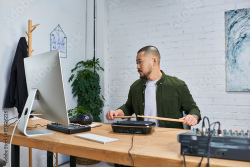 A handsome Asian man, wearing casual attire, sits in his studio and plays drums on an electronic drum pad. photo