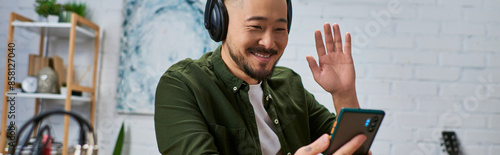 A handsome Asian man, wearing casual attire, sits in his studio, headphones on, waving to the camera. photo