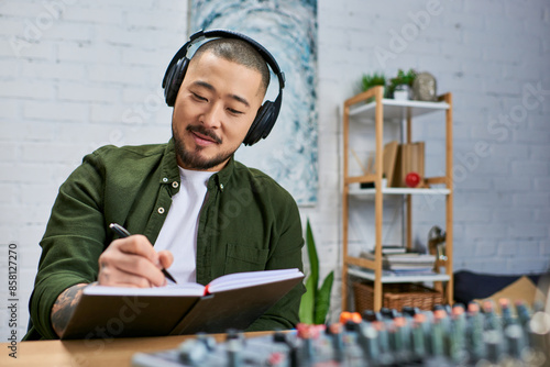 A young Asian man in casual wear writes in a notebook, wearing headphones in a music studio. photo