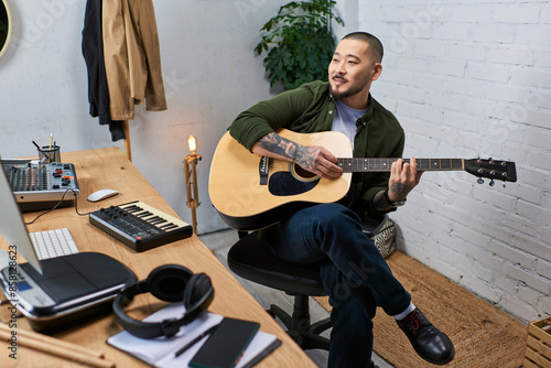 A handsome Asian man sits in his studio, playing an acoustic guitar.