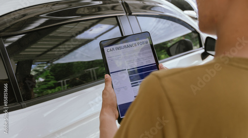 A young adult man, holding a smartphone, takes a photo of his dented car after an accident. An insurance agent with a clipboard examines the damage, preparing a claim report. photo