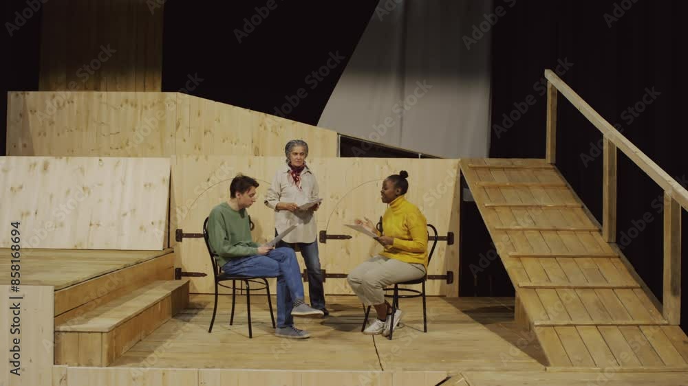 Young male and female drama class students sitting on chairs on stage, reading papers and practicing dialogue under control of female theatre director Young male and female drama class students sitting on chairs on stage, reading papers and practicing dialogue under control of female theatre director