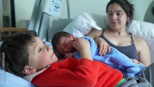 Older sibling lying on hospital bed with newborn baby, while mother watches lovingly in the background. tender family moment of bonding, warmth, and early sibling connection in a hospital setting