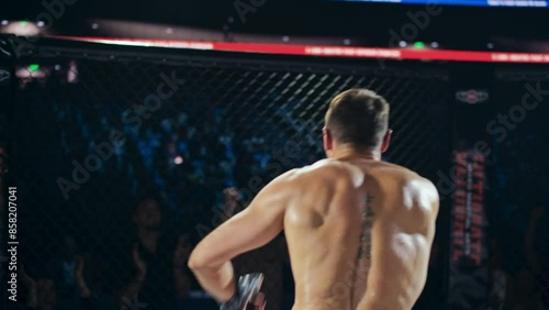 Referee Declares the Winner in a Tough MMA Bout Between Two Determined Male Fighters. Winning Fighter Raising His Hands in Triumph, Running and Climbing the Octagon Fence to Greet the Crowd photo