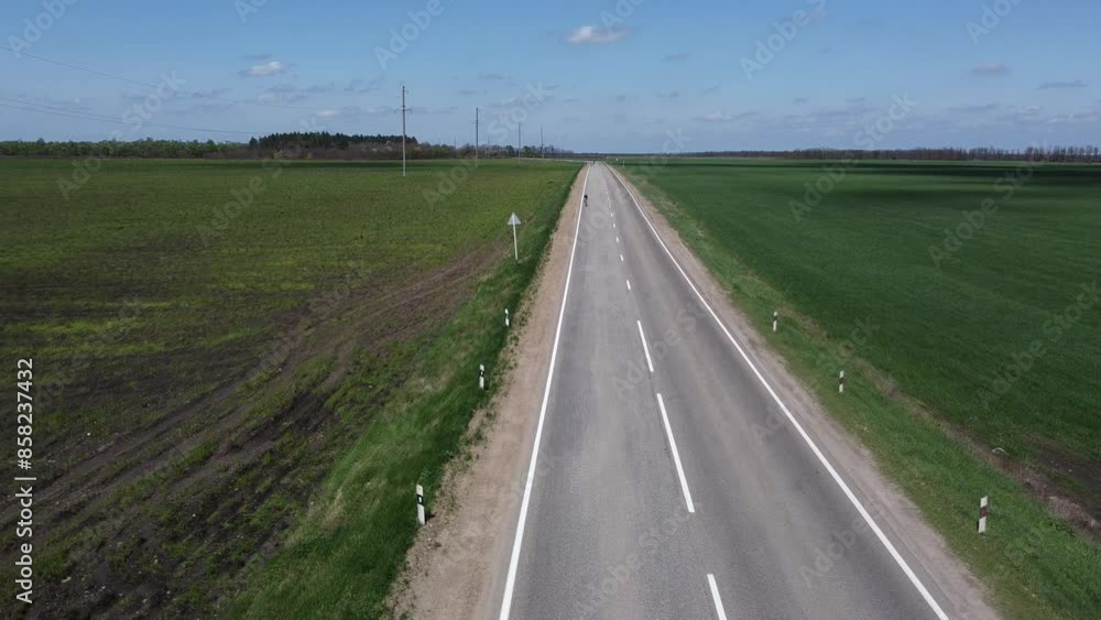 A lone cyclist is riding along the highway. A flat landscape. The time of the year is spring or summer.