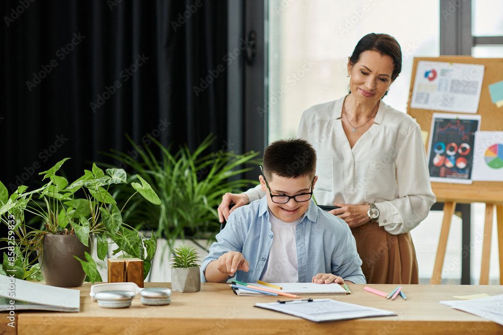 A mother stands behind her son with Down Syndrome as he works at a desk in an office setting.