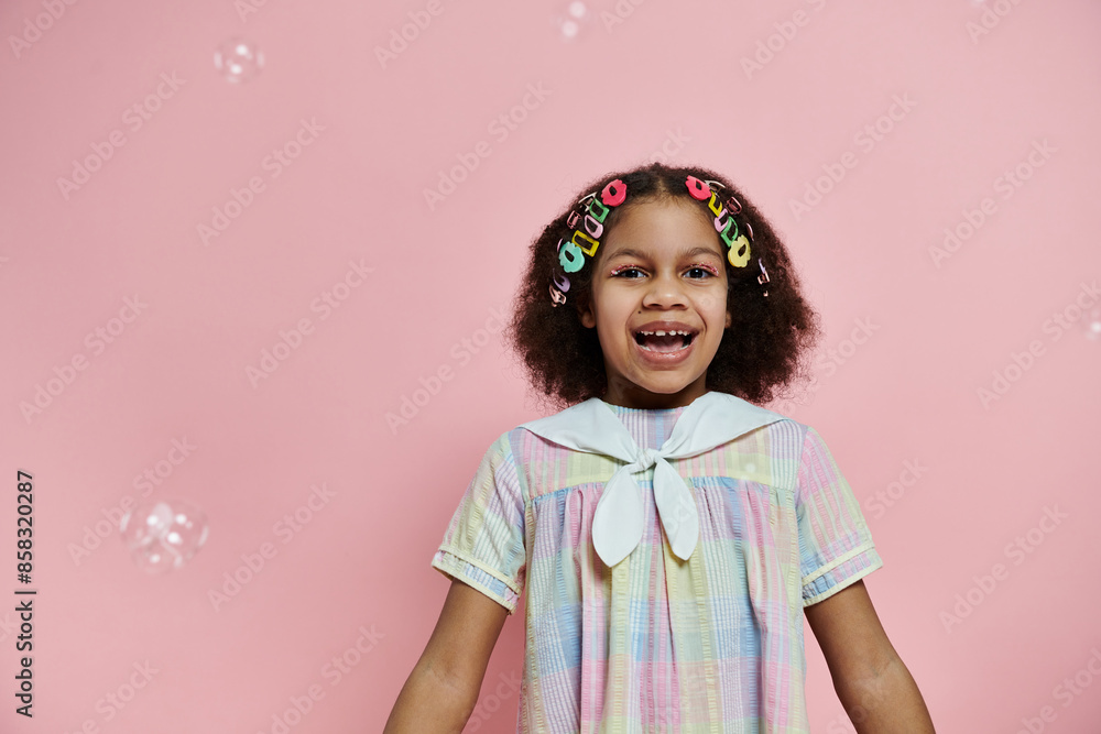 Young African American girl with hair clips in her hair smiles brightly in a pastel dress against a pink background. Young African American girl with hair clips in her hair smiles brightly in a pastel dress against a pink background.