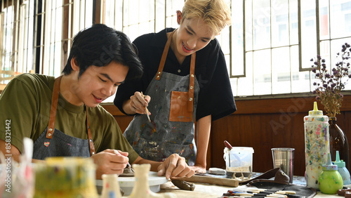 An art teacher assists a student in sculpting clay at a pottery wheel. Both are focused and smiling in a bright studio filled with art tools, paint supplies, and natural light from large windows