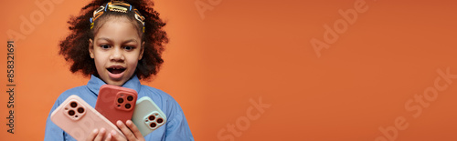 A young African American girl in a blue shirt stares in awe at a collection of colorful phone cases against an orange backdrop.