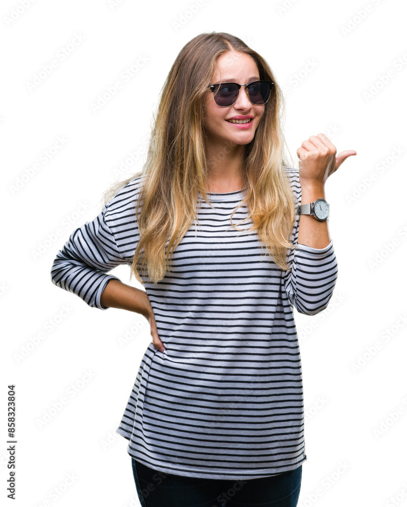 Young beautiful blonde woman wearing sunglasses over isolated background smiling with happy face looking and pointing to the side with thumb up. Young beautiful blonde woman wearing sunglasses over isolated background smiling with happy face looking and pointing to the side with thumb up.