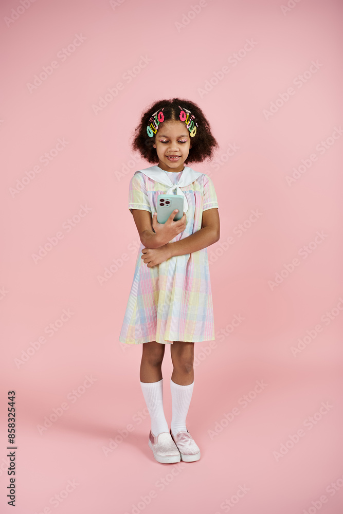A young African American girl with colorful hair clips stands in a cute dress on a pink background. A young African American girl with colorful hair clips stands in a cute dress on a pink background.