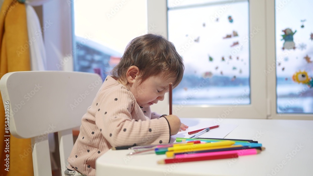 baby girl draws while sitting at a table by the window at home. happy family kid concept. baby daughter learns to draw with pencils on a sheet of paper indoors. development dream of fine motor skills baby girl draws while sitting at a table by the window at home. happy family kid concept. baby daughter learns to draw with pencils on a sheet of paper indoors. development dream of fine motor skills