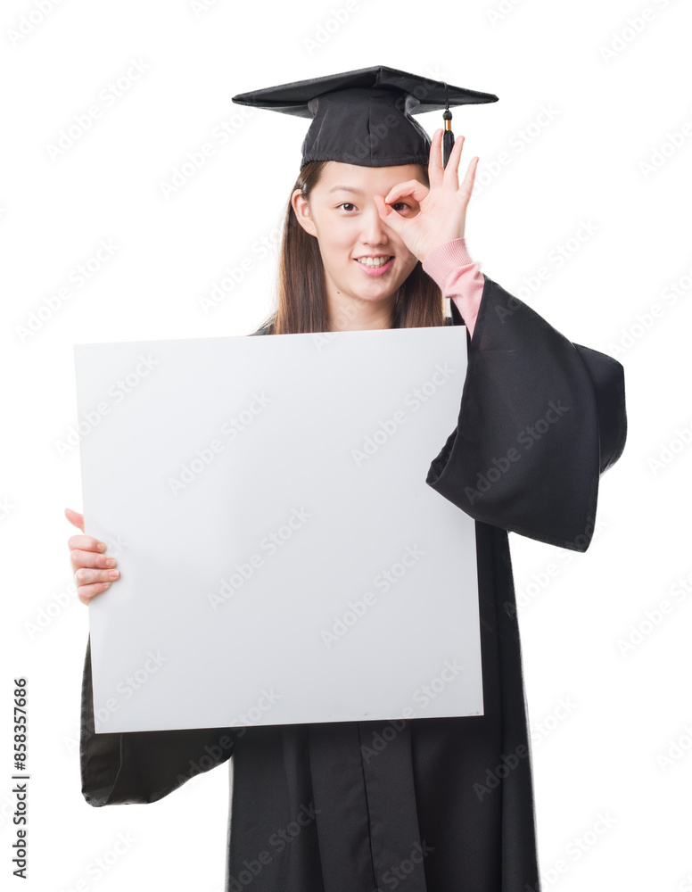 Young Chinese woman wearing graduate uniform holding banner with happy face smiling doing ok sign with hand on eye looking through fingers Young Chinese woman wearing graduate uniform holding banner with happy face smiling doing ok sign with hand on eye looking through fingers