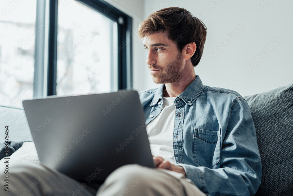 Handsome serious man sits on sofa with computer on laps, spend time at home web surfing information, use modern tech, make order on internet. Freelancing, telework, new software learning, tech concept Handsome serious man sits on sofa with computer on laps, spend time at home web surfing information, use modern tech, make order on internet. Freelancing, telework, new software learning, tech concept