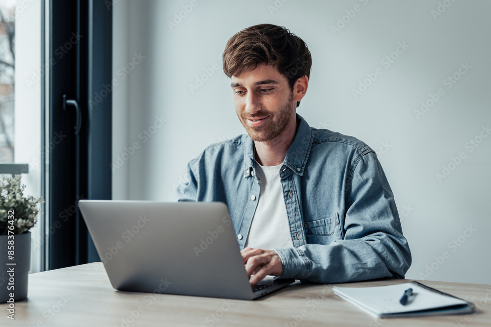 Side view handsome young businessman working with computer remotely, sitting at wooden table in office. Pleasant happy man communicating in social network, searching information online.. Side view handsome young businessman working with computer remotely, sitting at wooden table in office. Pleasant happy man communicating in social network, searching information online..