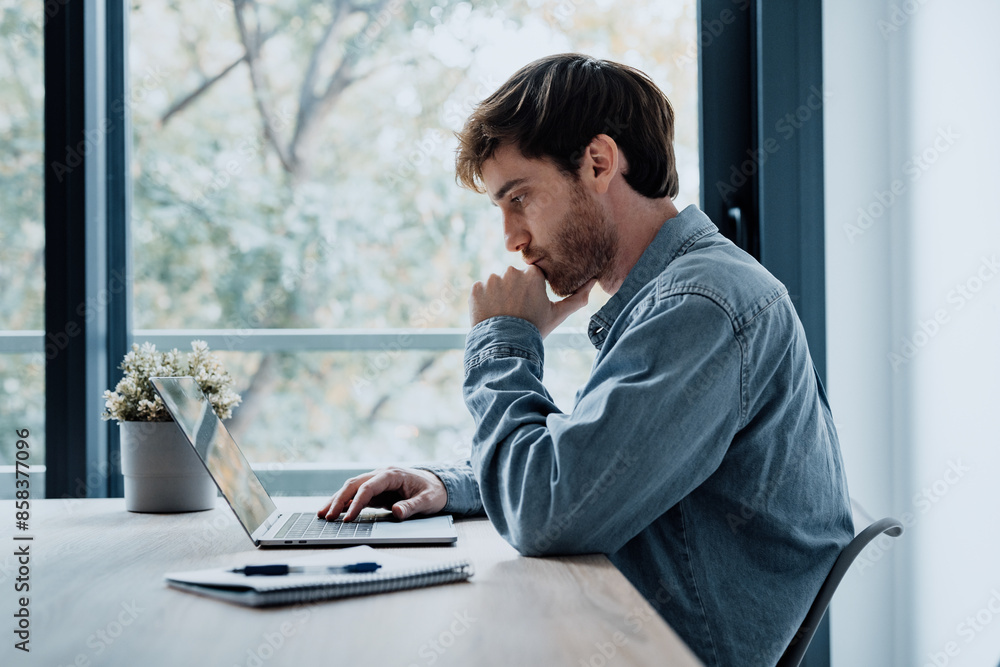 Thoughtful businessman touching chin, pondering ideas or strategy, sitting at wooden work desk with laptop, freelancer working on online project, student preparing for exam at home. Thoughtful businessman touching chin, pondering ideas or strategy, sitting at wooden work desk with laptop, freelancer working on online project, student preparing for exam at home.