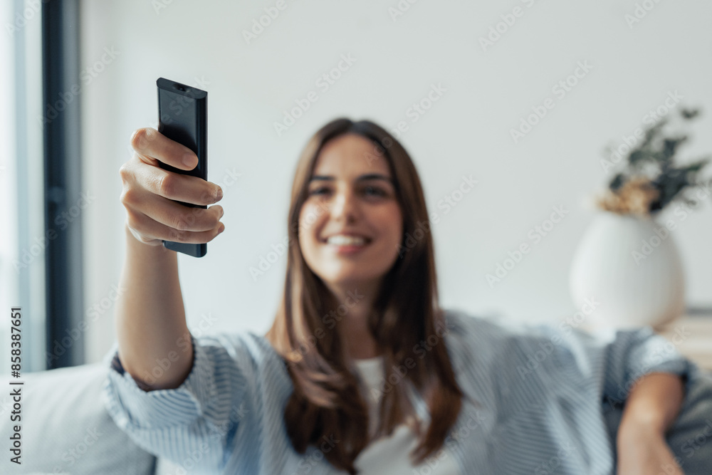 Switching on. Happy millennial female user sit on sofa at living room controlling smart tv set air conditioner home theater system. Focus on infrared remote control device in young woman customer hand