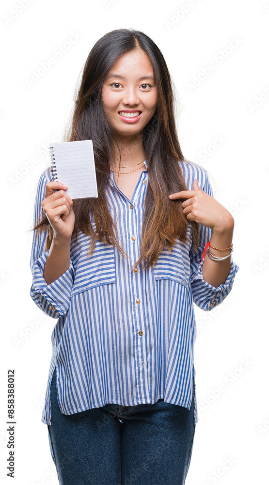 Young asian woman holding notebook over isolated background with surprise face pointing finger to himself Young asian woman holding notebook over isolated background with surprise face pointing finger to himself