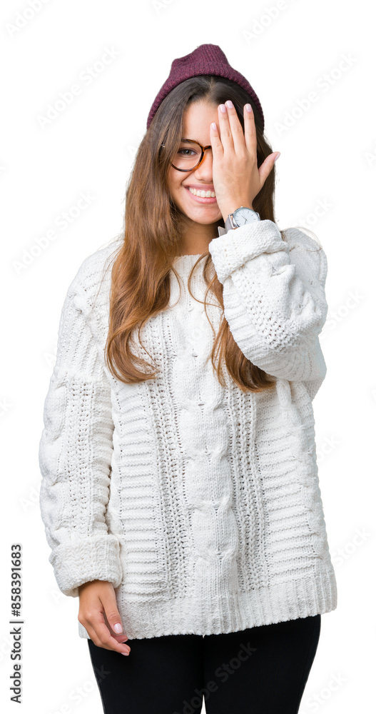 Young beautiful brunette hipster woman wearing glasses and winter hat over isolated background covering one eye with hand with confident smile on face and surprise emotion. Young beautiful brunette hipster woman wearing glasses and winter hat over isolated background covering one eye with hand with confident smile on face and surprise emotion.