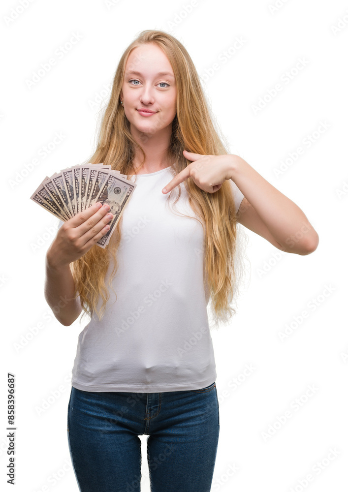 Blonde teenager woman holding dollars with surprise face pointing finger to himself Blonde teenager woman holding dollars with surprise face pointing finger to himself