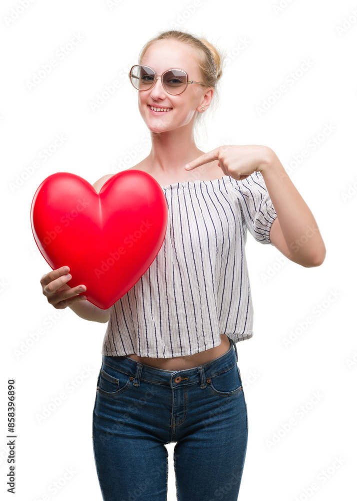 Blonde teenager woman holding red heart very happy pointing with hand and finger Blonde teenager woman holding red heart very happy pointing with hand and finger