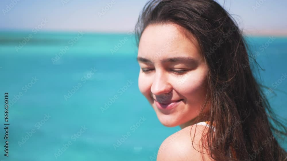 A young attractive girl looks at the camera and smiles against the background of the blue ocean. Young millennial girl enjoying a wonderful holiday at sea. A young attractive girl looks at the camera and smiles against the background of the blue ocean. Young millennial girl enjoying a wonderful holiday at sea.