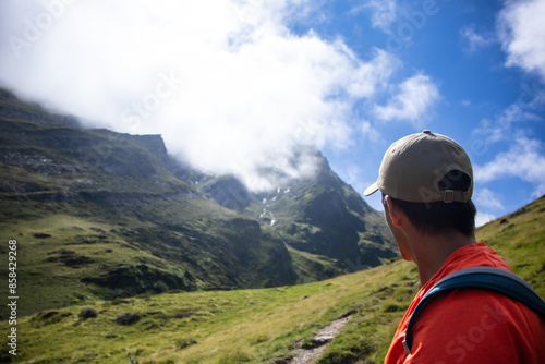 Rear view of a young man in a cap and trekking outfit looking towards the top of the mountain on a sunny summer day with some clouds