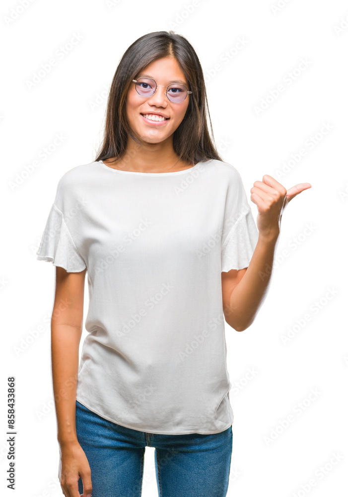 Young asian woman wearing glasses over isolated background smiling with happy face looking and pointing to the side with thumb up. Young asian woman wearing glasses over isolated background smiling with happy face looking and pointing to the side with thumb up.