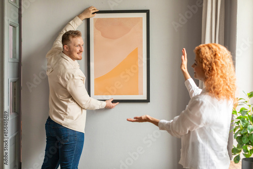 Bilde på lerret Young spouses arranging new flat, hanging up painting on wall