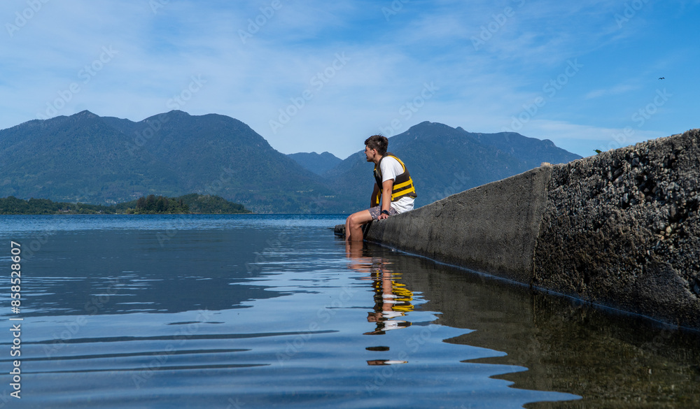 Picture-perfect scene of a young man seated on the lake dock, taking in the stunning vista during a delightful summer afternoon. Picture-perfect scene of a young man seated on the lake dock, taking in the stunning vista during a delightful summer afternoon.