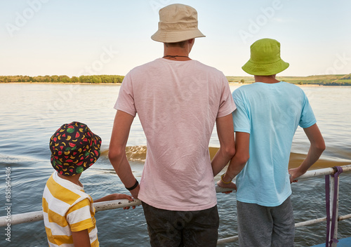 Three boys in colorful bucket hats and casual t-shirts stand on a railing by the water, looking at the scenic view of the river and distant trees on a sunny day. photo