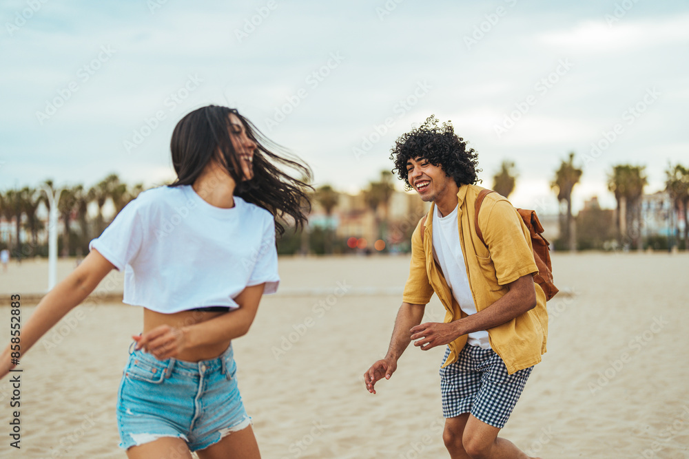 A young Hispanic man and woman laugh while playfully running on the beach, dressed in casual summer clothes with a backpack suggesting a day trip. A young Hispanic man and woman laugh while playfully running on the beach, dressed in casual summer clothes with a backpack suggesting a day trip.