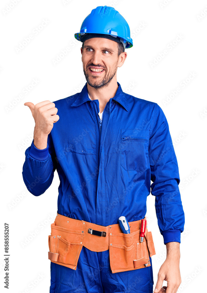 Young handsome man wearing worker uniform and hardhat smiling with happy face looking and pointing to the side with thumb up. Young handsome man wearing worker uniform and hardhat smiling with happy face looking and pointing to the side with thumb up.