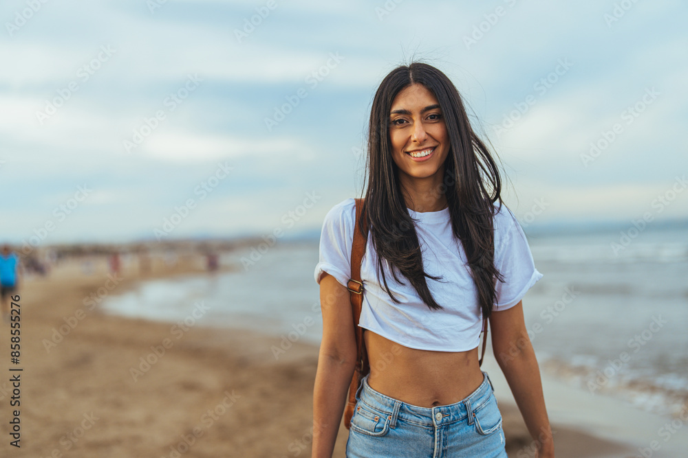 A smiling young Hispanic woman strolls along the beach, dressed casually in a white tee and denim shorts, embodying the leisure of a coastal setting. A smiling young Hispanic woman strolls along the beach, dressed casually in a white tee and denim shorts, embodying the leisure of a coastal setting.