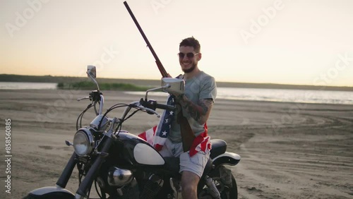 A man in sunglasses wripped in american flag sitting on motorcycle and rifle at sunset beach, embodying adventure, freedom, and a rugged, macho attitude in a scenic landscape photo