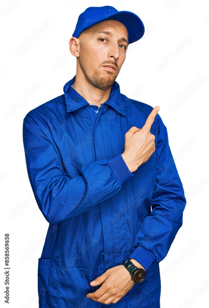 Bald man with beard wearing builder jumpsuit uniform pointing with hand finger to the side showing advertisement, serious and calm face Bald man with beard wearing builder jumpsuit uniform pointing with hand finger to the side showing advertisement, serious and calm face