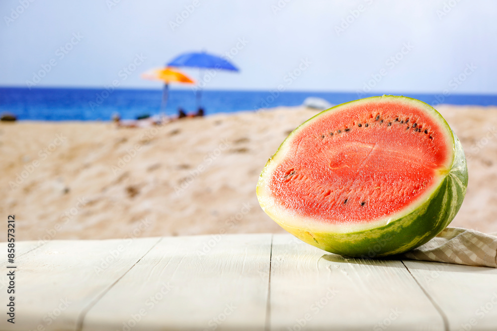 Half watermelon on white table top and blur background of sandy beach and sea and blue sky. Sun umbrellas in distance. Empty space for products and food. Half watermelon on white table top and blur background of sandy beach and sea and blue sky. Sun umbrellas in distance. Empty space for products and food.