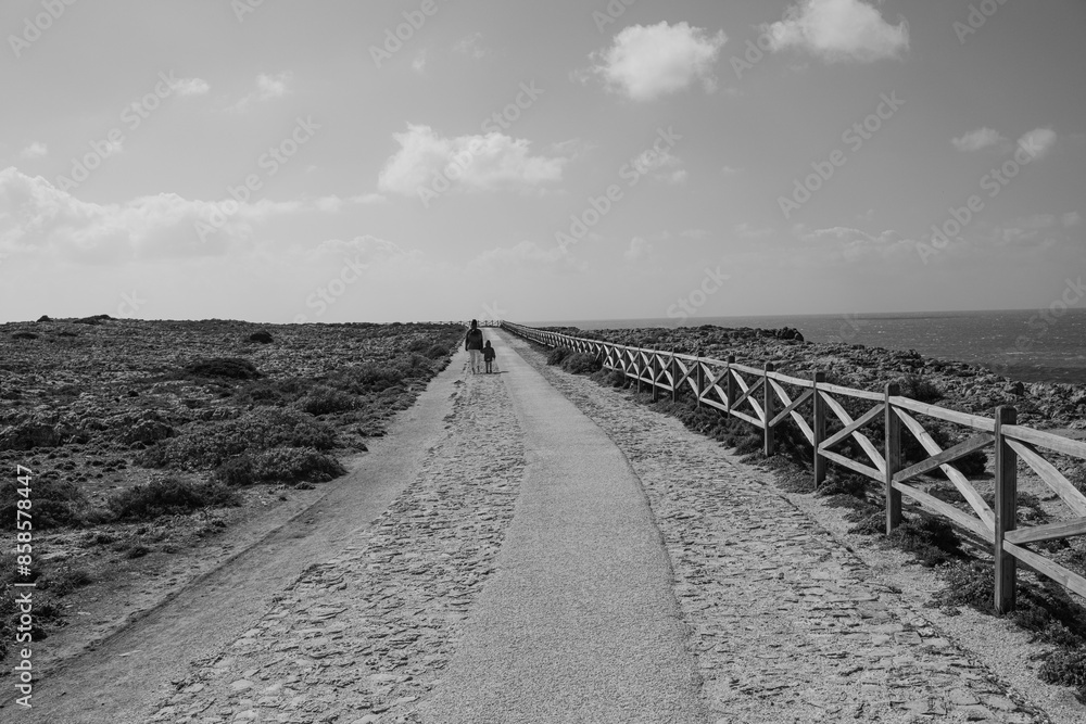 Fototapeta premium A mother and child walk along a path along the ocean. Mom and daughter in jackets walk along the road by the ocean in sunny and windy weather