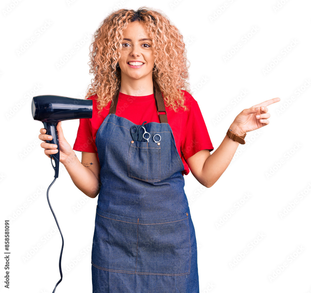 Young blonde woman with curly hair wearing hairdresser apron and holding dryer blow smiling happy pointing with hand and finger to the side Young blonde woman with curly hair wearing hairdresser apron and holding dryer blow smiling happy pointing with hand and finger to the side