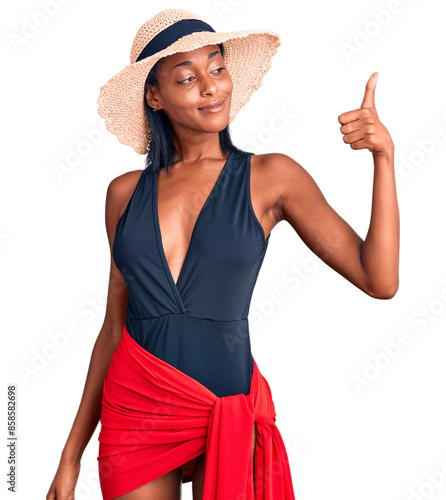 Young african american woman wearing swimsuit and summer hat looking proud, smiling doing thumbs up gesture to the side photo