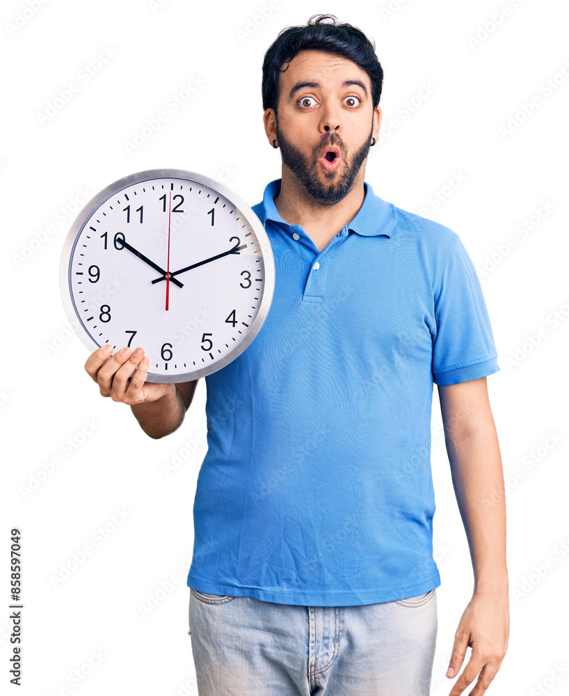 Young hispanic man holding big clock scared and amazed with open mouth for surprise, disbelief face Young hispanic man holding big clock scared and amazed with open mouth for surprise, disbelief face