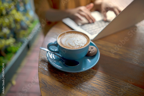 A person is typing on a laptop while a blue coffee cup with a cookie sits on the