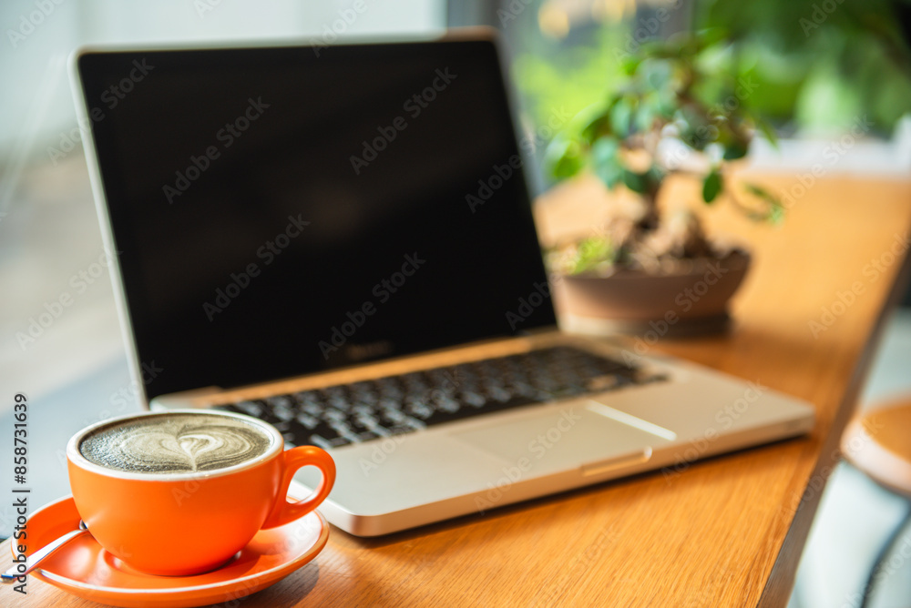 Work area with laptop Orange coffee with latte art and small potted plants The focus is on the coffee cup. with a laptop and trees in the background In a coffee shop or home office Work area with laptop Orange coffee with latte art and small potted plants The focus is on the coffee cup. with a laptop and trees in the background In a coffee shop or home office
