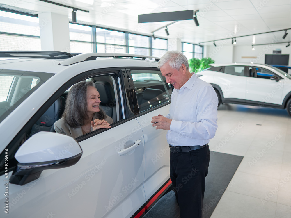 An elderly Caucasian woman is sitting in a new car. Her husband is standing nearby.
