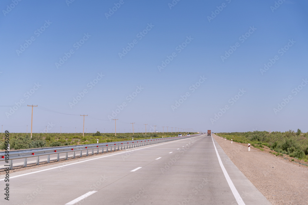 A long, empty highway with a truck driving down it. The sky is clear and blue, and there are no other vehicles on the road A long, empty highway with a truck driving down it. The sky is clear and blue, and there are no other vehicles on the road