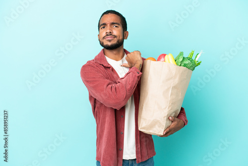 Young latin man holding a grocery shopping bag isolated on blue background proud and self-satisfied