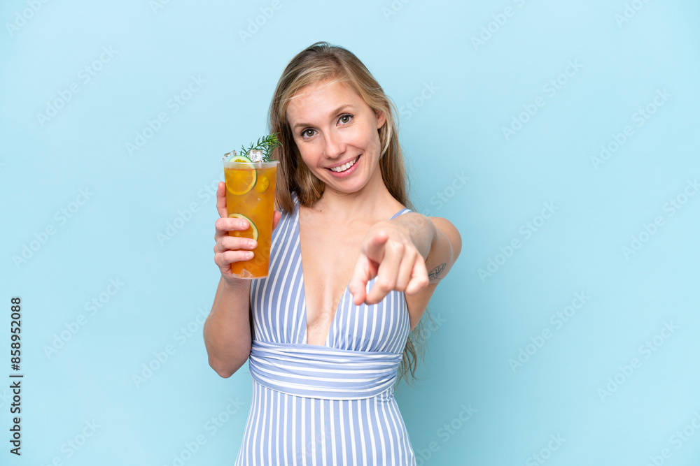 Young blonde woman wearing swimsuit holding a cocktail isolated on blue background points finger at you with a confident expression Young blonde woman wearing swimsuit holding a cocktail isolated on blue background points finger at you with a confident expression