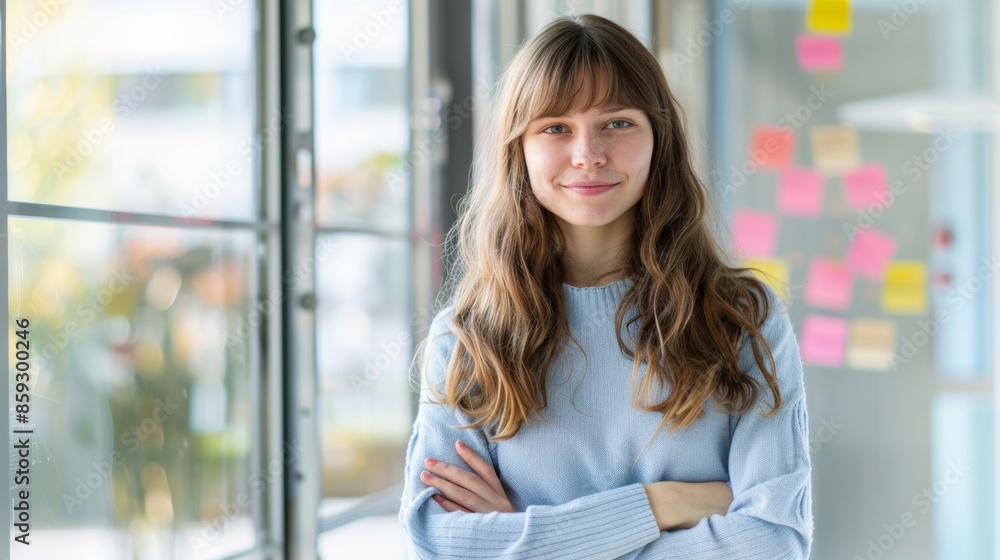 Smiling young woman standing with crossed arms in a bright, modern office setting, with charts and post-it notes on the window behind her Smiling young woman standing with crossed arms in a bright, modern office setting, with charts and post-it notes on the window behind her
