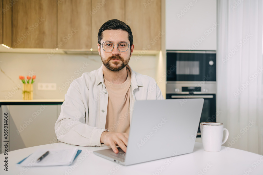 Portrait of cheerful bearded young man using laptop sitting at table in kitchen room with modern interior, looking at camera remote working from home. Portrait of cheerful bearded young man using laptop sitting at table in kitchen room with modern interior, looking at camera remote working from home.