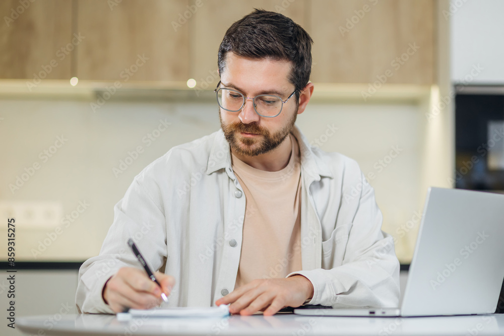 Young man student prepare research essay at modern kitchen surf internet use laptop write up information to paper report by hand. Focused man freelancer work from home make list of data from screen Young man student prepare research essay at modern kitchen surf internet use laptop write up information to paper report by hand. Focused man freelancer work from home make list of data from screen